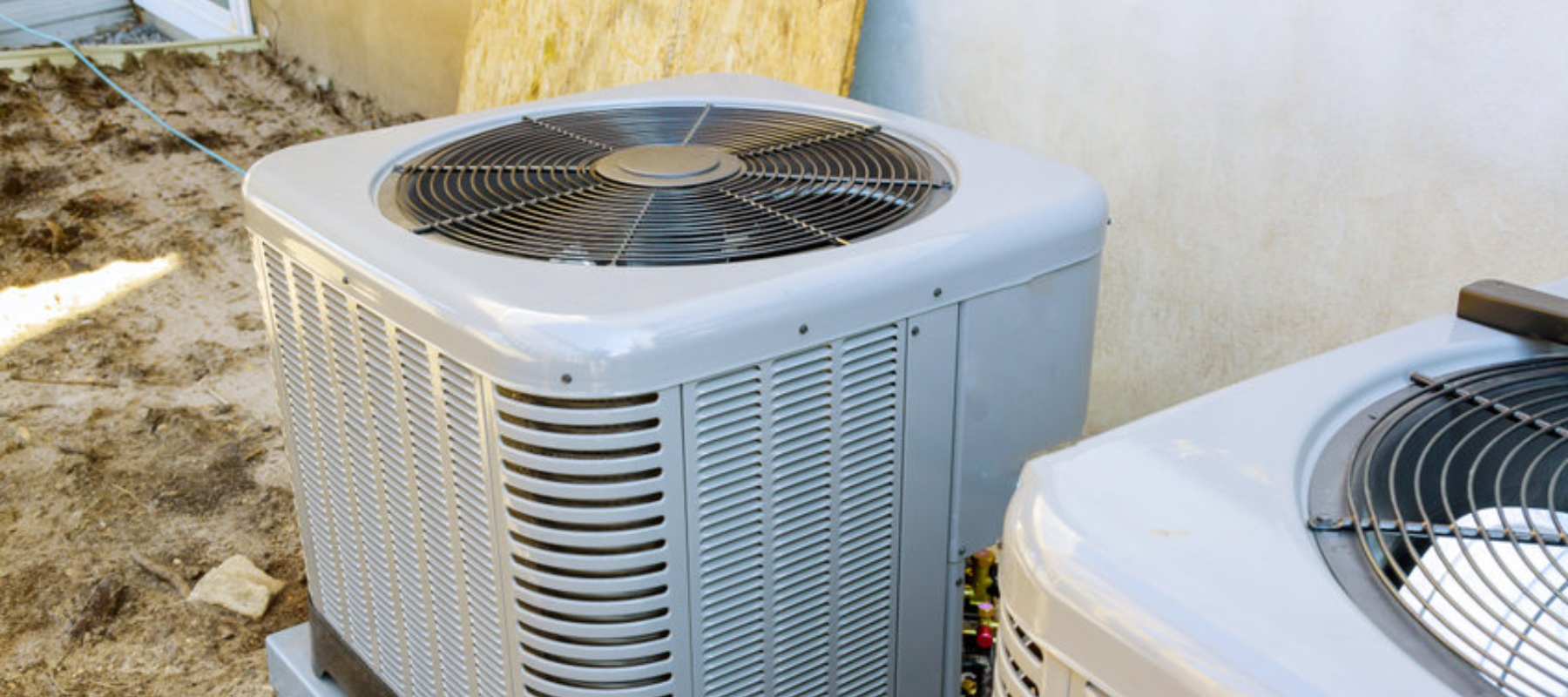 Two outdoor HVAC condenser units sitting on bare ground next to a wall.