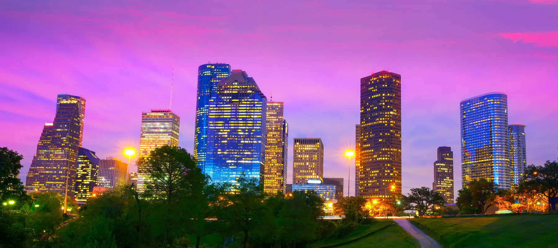 Downtown city skyline at sunset with tall buildings illuminated, trees in the foreground, and a pink and purple sky overhead.