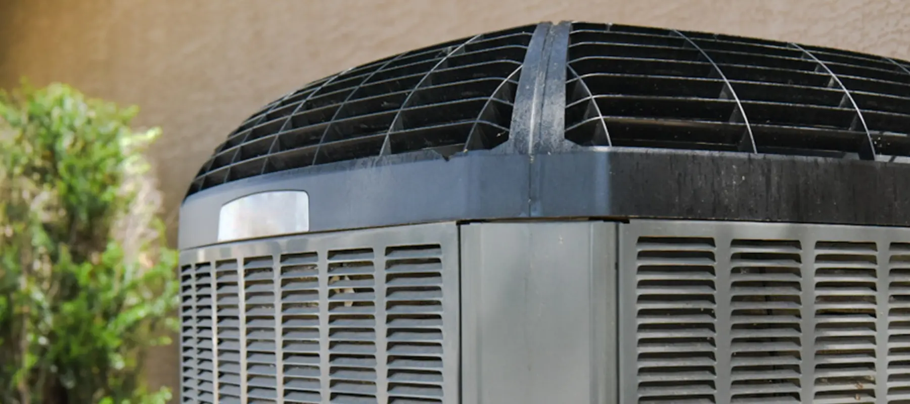 A close-up view of a gray outdoor air conditioning unit with black grill vents, positioned near a tan wall and some green plants.