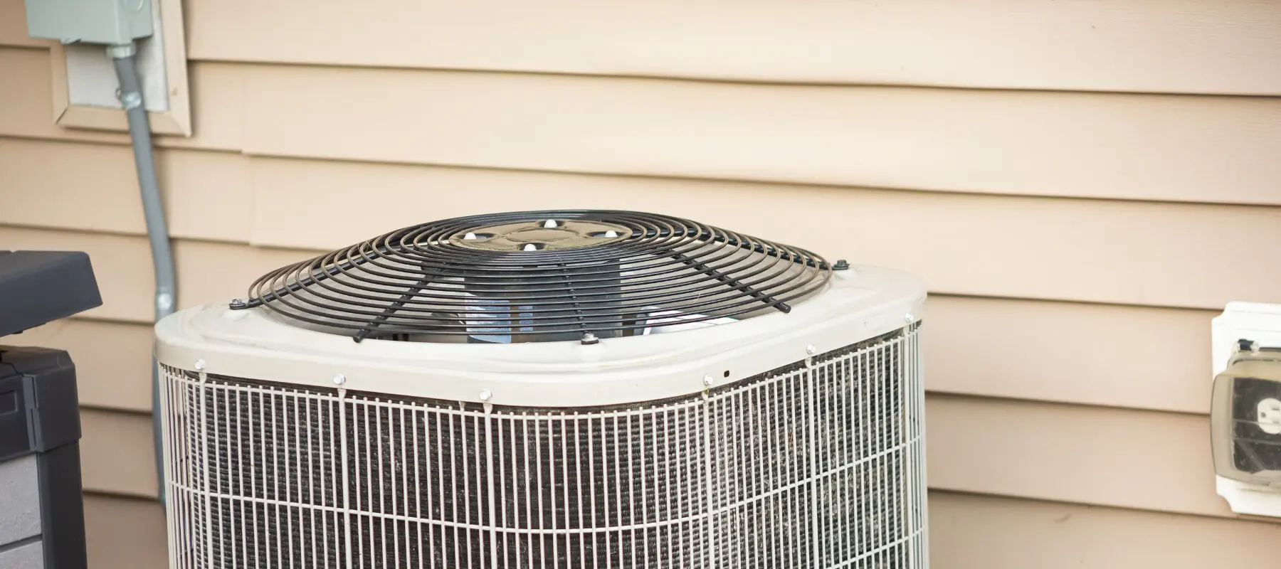 Outdoor air conditioning unit next to a beige vinyl-sided house wall, with a black fan cover on top and visible coils on the sides. Electrical connections are seen nearby.